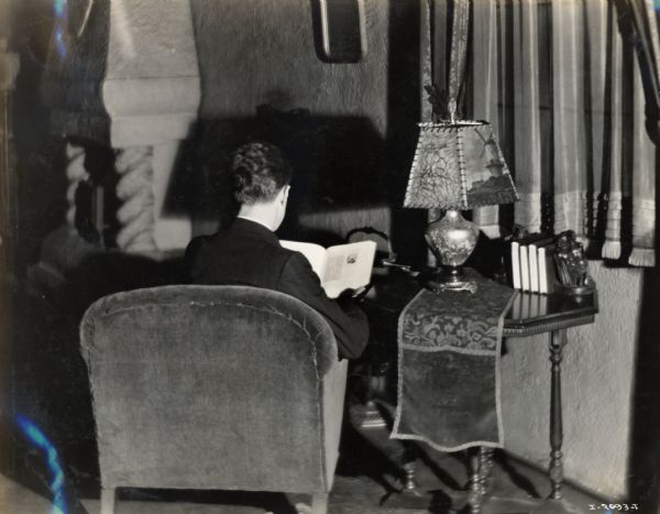 A man reads in a chair, with his back to the camera. His book rests on a table near a lamp with a decorated shade.