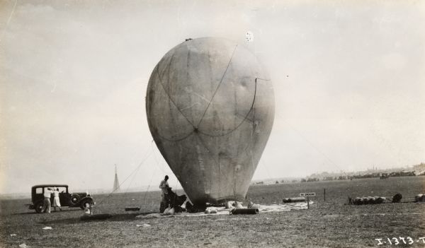 A balloon sits in a field at the National Air Show, held at Curtiss-Reynolds Airport. Two people stand near an automobile on the left, and on the right near the balloon is a sign near gas tanks that reads: "No Smoking". In the background there appears to be a crowd on a grandstand.