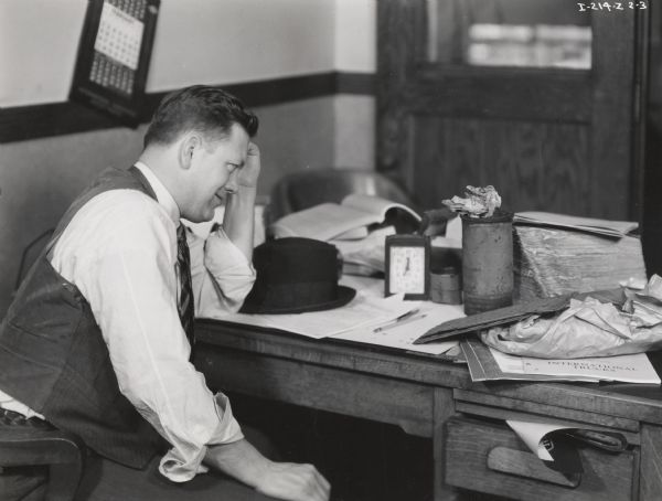 A man at the Number 1 Motor Truck Station sits at a unorganized desk. Possibly a photograph from an instructional film about proper International Harvester salesmanship. This photograph is an illustration of poor salesmanship.