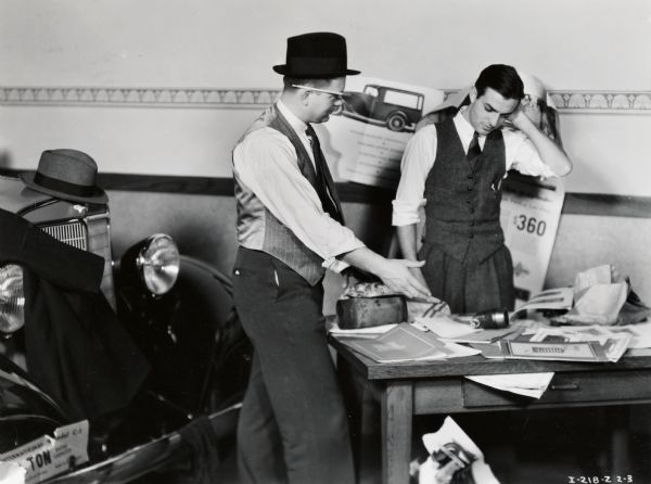 Two men stand over a disorganized desk on a salesroom floor at the Number 1 Motor Truck Station. Possibly a photograph from an instructional film about proper International Harvester salesmanship. This photograph is an illustration of poor salesmanship.