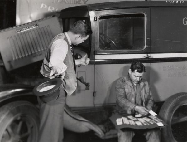 Two men in the Number 1 Motor Truck Station shop. A salesman stands over a mechanic, who appears to be playing a game of solitaire. Possibly a photograph from an instructional film about proper International Harvester salesmanship. This photograph is an illustration of poor salesmanship.
