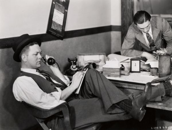 Two men, one talking on the telephone with his feet on the desk and another reading paperwork, work at a disorganized desk. Possibly a photograph from an instructional film about proper International Harvester salesmanship. This photograph is an illustration of poor salesmanship.