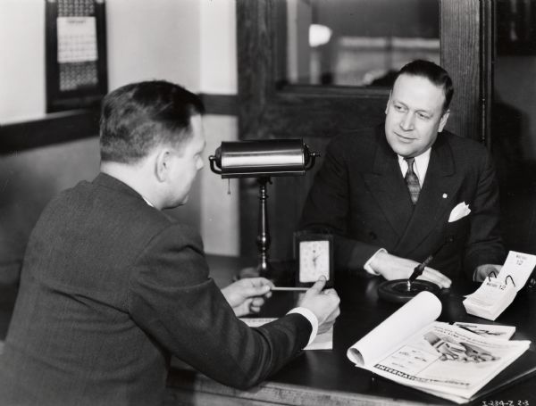 Two men converse in a tidy office. Possibly a photograph from an instructional film about proper International Harvester salesmanship. This photograph is an illustration of good salesmanship.