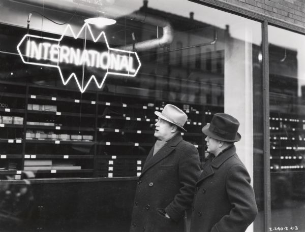 Two well-dressed men stand outside and look at a neon International triple diamond truck logo displayed in a window. Possibly a photograph from an instructional film about proper International Harvester salesmanship.