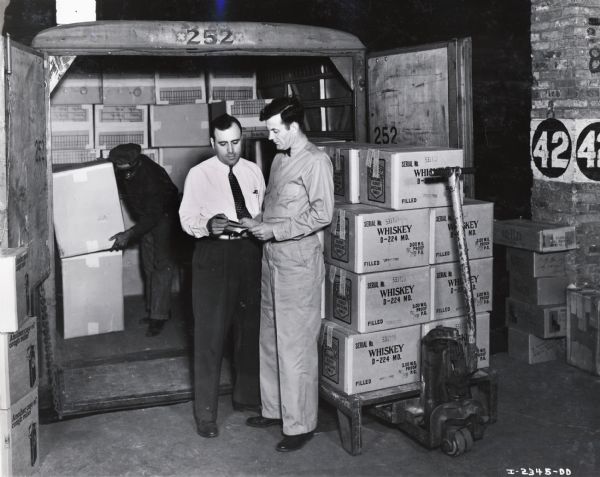 Two men look at paperwork while another moves boxes with a truck. Original caption reads: "David Davidson, in white shirt, and loading foreman Louis Holter, Davidson Transfer and Storage Co."