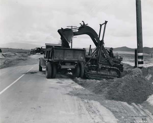 International TD-9 crawler tractor (TracTracTor) lifts dirt into a dump truck. Original caption reads: "An International TD-9 Diesel TracTracTor equipped with 3/4-yard hydraulically operated front-end Be-Ge shovel and owned by Ed Waters of Los Angeles, was recently rushed down to Highway No. 395, some five miles south of Elsinore, to remove from 700 to 800 yards of flood debris. Material was first moved to side of highway and then quickly loaded into trucks, as shown."