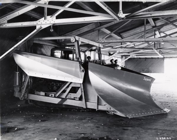 A large plow is attached to a crawler tractor. Original caption reads: "International TD-18 Diesel wide-tread TracTracTor provided with cab and complete winter equipment owned by the Town of Arlington, Massachusetts. Operation of the tractor is under the control of the Board of Public Works, Town Hall, Arlington, and consists of three members, Willard A. Wallace, (chairman), Loran W. Marsh, and James R. Smith."