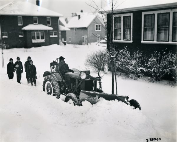 A man operating an International I-12 tractor with a snowplow to plow a sidewalk. A group of children are  walking behind the snowplow.