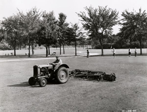A man mows a golf course with an International I-12 tractor owned by the City of Davenport. A group of women are playing golf in the background.