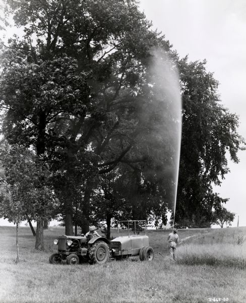 Two men are operating an International I-14 tractor and a beam sprayer. One man is sitting on the tractor while the other man is standing and operating the sprayer.