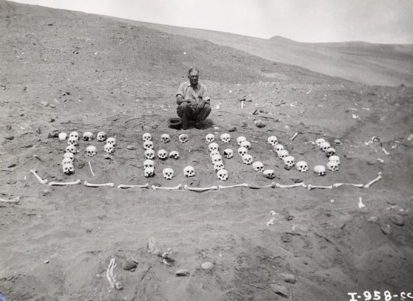 A man crouches behind an arrangement of human skulls and bones that spell "Peru". Caption on photograph reads: "Illustrations for article on the TracTracTor in Peru, appearing in June 1939 <i>International Power</i>."