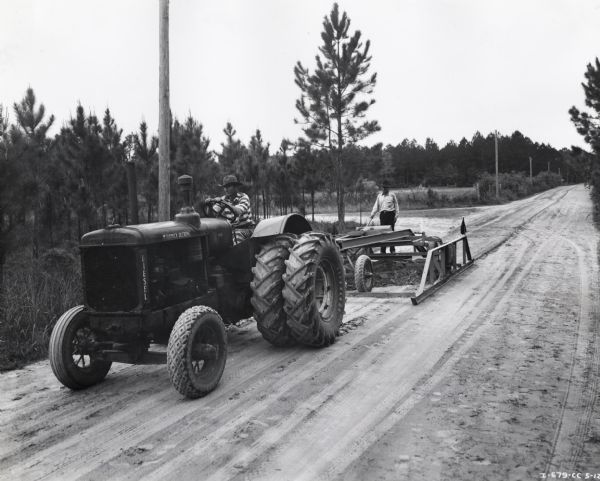 Two men operate an International Harvester Diesel Tractor. Original caption reads: "International Diesel tractor with dual pneumatic tires and new type of Allen 10-foot heavy-duty maintainer owned by Nassau County, Florida, and used for maintaining roads at a speed of 7 to 10 miles per hour. New features of the maintainer include hydraulic pressure to keep the blade from shattering, and side runners which serve as depth gauges. Once over with this maintainer does same quality of work as two to four times over with a 12-foot maintainer which was formerly used by the county and which did not have these features. The Diesel tractor uses only 1 1/2 gallons of 7-cent fuel an hour and has operated approximately 5,000 hours with a repair expense of only $15."