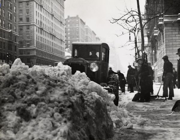 A man plows snow with an International I-30(?) tractor on a New York City street. Men with shovels are standing on the sidewalk.