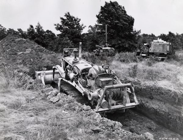 Man operates an International T-40 crawler tractor. Original caption reads: "International T-40 TracTracTor equipped with Bucyrus-Erie bulldozer and Tulsa winch owned by Helmerich and Payne Inc., drilling contractors of 1702 Philtower Building, Tulsa, Oklahoma, building slush pit in Salem field. Pit is 75-feet long, 5-feet deep and 9-feet 2-inches wide. Soil is gumbo clay. A "complete set" (two slush pits this size and also a reserve pit) can be dug in 2 hours, but usual time is about 3 hours. This outfit has dug 70 "complete sets" in Illinois fields. Operator rollers on other makes of crawler tractors, and that for speed, ease of handling, and low cost, "there is nothing like International and Bucyrus-Erie" for slush pits. The winch has been used time and time again during the past winter for pulling trucks and other heavy equipment out of the mud."