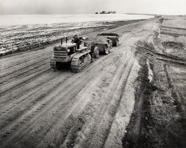 Elevated view of a man operating a TD-18 crawler tractor to level a road in the wintertime. There is a farm in the background. Original caption reads: "TD-18 and Isaacson 6 yards rope working on frozen ground preparing for the construction of a new state highway near Ritzville, Washington."