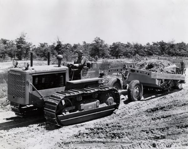 A man operates an International TD-18 crawler tractor (TracTracTor) and a Bucyrus-Erie scraper. Original caption reads: "TD-18 TracTracTor and Bucyrus-Erie 4.5-yard 4-wheel scraper owned by A.F. Hartigan and Co., 5614 Dorchester Ave., Chicago. In use at site of new city of Gary sewage disposal plant where 200,000 cubic yards of dunes sand are being moved. Total contract, $5,000,000."