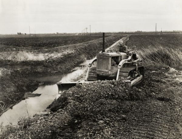 A man operates an International TD-35 TracTracTor (crawler tractor) to create a slope for a levee. Original caption reads: "A TD-35 TracTracTor with 10 foot Bucyrus-Erie bullgrader sloping the levee on the Sacramento River to prevent flood waters from washing away the levee."