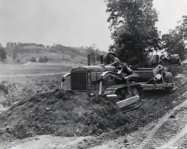 Man operating an International TD-40 TracTracTor (crawler tractor) with a bulldozer and two wheel scraper attached. The tractor is possibly leveling dirt to make way for a new road.