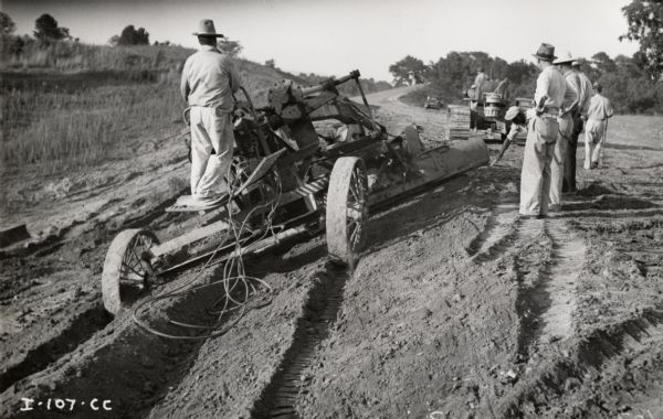 A man operates a grader pulled by an International TD-18 TracTracTor (crawler tractor) as other men observe. Original caption reads: "Leaning wheel grader operated by TD-18. Taken by John Erskins-Gas Power Engineering."