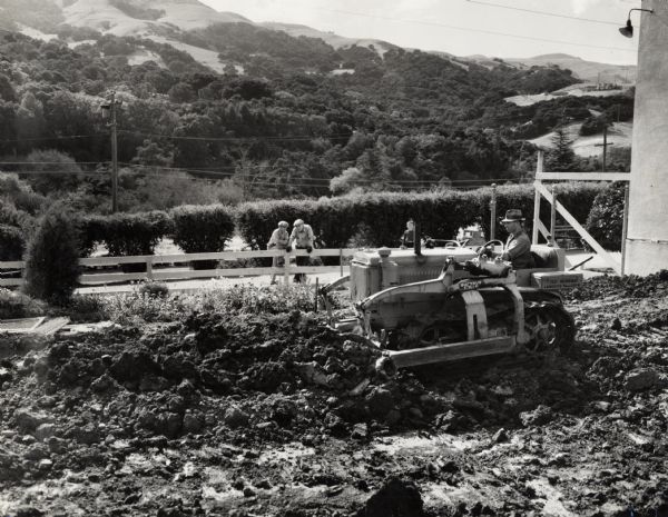 A man operates a crawler tractor to level an area of dirt while two men near a fence observe. Original caption reads: "A T-20 TracTracTor and Bucyrus-Erie bulldozer, owned by Ted Oliveira. Lafayette, Cal. Excavating basement for new addition to the Corinda(?) Public School."