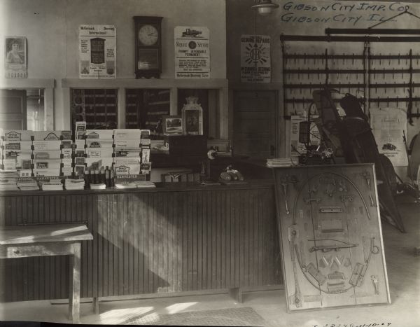 Customer service counter at Gibson City Implement Company, an International Harvester agricultural equipment dealership. A display board of parts is leaning against a counter with pamphlets, a phone, corn, and other objects. A clock, posters, and equipment are along the wall on the right.