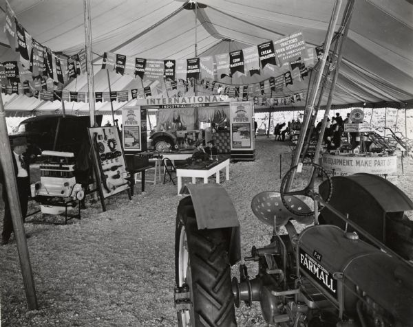 People browse through an International Harvester display tent, possibly at the Tennessee Valley Fair. The tent features displays of International Harvester parts and machinery, including a Farmall F-14 tractor, an I-12 industrial tractor, an engine, binder twine, and an International truck.