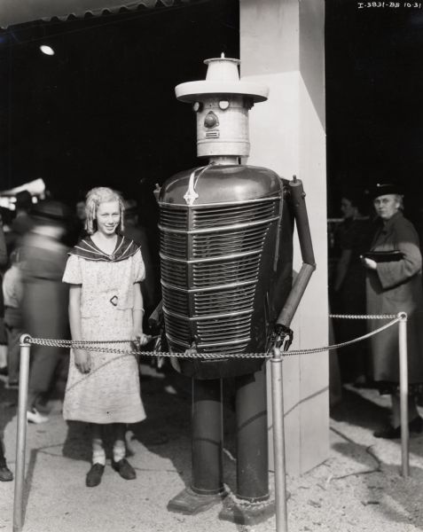 A young girl standing next to "Harvey Harvester," a talking robot made of parts from International Harvester machines, including the grill of an International truck. The robot was likely on display at the Waterloo Dairy Cattle Congress or at the Iowa State Fair. "Harvey Harvester" was a precursor to "Tracto" the "talking robot," which IH introduced in 1960.