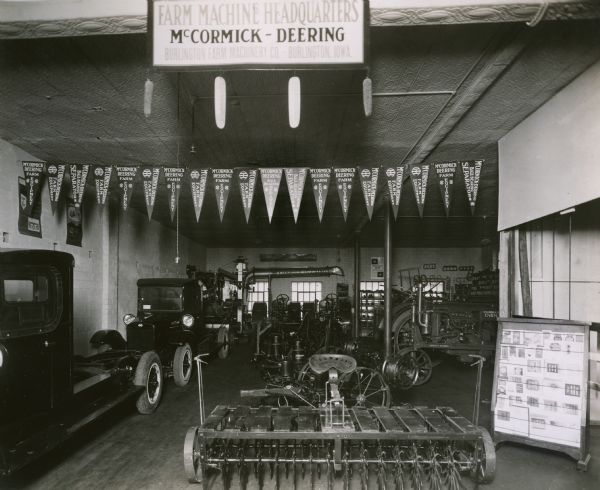 Trucks, tractors, and farm equipment in the showroom of the Burlington Farm Machinery Company, an International Harvester dealership. Advertising pennants hang from the ceiling. Brochures and other literature are on a rack on the right.