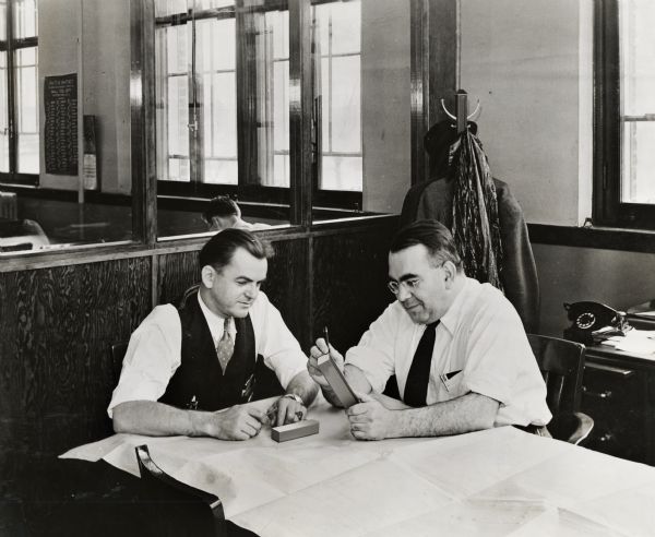 Mechanical engineer Alex Gabay (right) and his assistant J.E. Lass (left) at an International Harvester war production factory (20 mm gun plant?). The men have a tool layout drawing on the table in front of them.