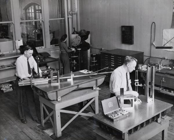 Factory inspectors at International Harvester's 20 mm gun plant. A woman also stands in the background using a piece of equipment. Original caption reads: "Some of the $175,000 worth of equipment for checking gauges and other work in the layout room of the Inspection Department is shown here. Of the two men inspectors at the surface plates, one is examining a gun tube and the other the unit of a breech block."