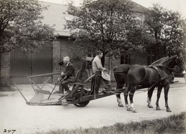 Two men use horses and a rake to demonstrate a McCormick reaper of 1849 in Copenhagen, Denmark. A building and trees are in the background.