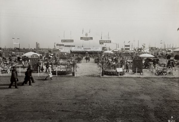 Display area of International Harvester's Neuss branch house at the Exhibition of the German Agricultural Society, Cologne, Germany, (May 27-June 1). Equipment and tractors are on display, with a path leading to a tent. There is a clock tower made out of twine in front of the tent. Smokestacks, signs, and a tall building are in the background.  The translation of the sign on the roof is: "Telegram from Chicago: On the 13th of May the 600,000th I.H.C. tractor came off of the endless chain [assembly line?]," and the sign hanging from the roof says: "Good Equipment Makes a Good Farmer Better."
