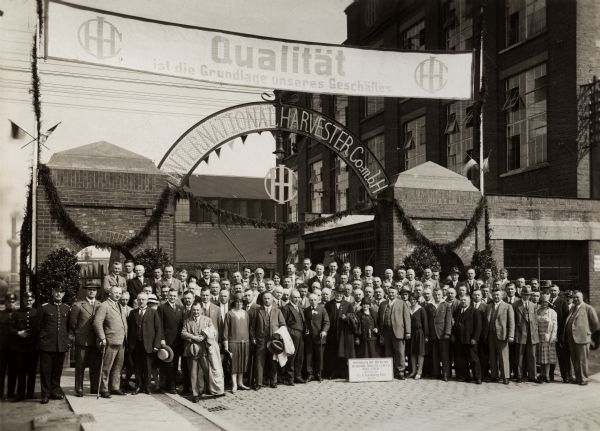 A crowd at the front gate of International Harvester's Neuss Works.  During the German Agricultural Society Show in Cologne (May 27-June 1, 1930) excursions of visitors consisting of agents and farmers were made daily to the factory. The original caption reads: "A Happy Family before the Gates on the Morning of May 31, 1930". A large group of people stands with a sign, some in hats.  Two men in uniform stand to the left side.