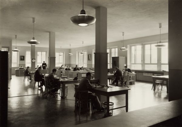 Men and women sitting at desks in a large, open office at International Harvester's Neuss branch in Germany. One man in the back is standing and reading a paper. Large light fixtures hang from the ceiling. Light comes in through the large windows.