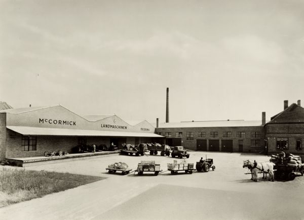 Exterior of International Harvester's Magdeburg branch house in Germany, with trucks, a tractor pulling three loaded carts, and a horse-drawn wagon piled with sacks. On the building above the platform is written: "McCormick / Landmashinen / Deering."