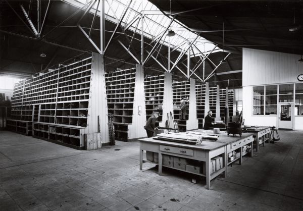 Men in a work room with parts bins at International Harvester's Hamburg branch house in Germany. An enclosed office is on the right, with a clock above the entrance. A skylight provides light into the room. A third man is standing on the second level of shelves in the background.