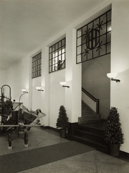 Staircase off of a showroom at International Harvester's branch house at Breslau, Germany. Ironwork over the staircase includes an IH logo. A woman, seen through the decorative grating, is on the staircase. Original caption reads: "Maschinen-Ausstellungsraum - Aufgang zum Obereschoss."