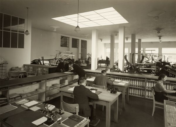 Office workers at desks next to a showroom at International Harvester's branch house in Breslau, Germany. A skylight illuminates reapers, grain binders and other agricultural machinery on display.