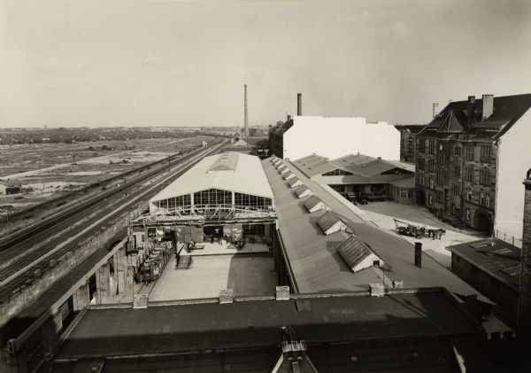 Elevated view of several buildings with railroad tracks at International Harvester's Berlin-Tempelhof branch house in Germany. Trucks and a horse-drawn wagon are on the right. Original caption includes the text: "Maschinen - und Ersatzteil-Lagerhauser mit Hofen und Gleisanschluss."