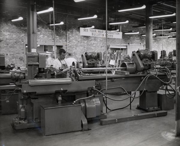 A factory worker operates a grinder at International Harvester's 20 millimeter gun plant. Original caption reads: "The Harvester gun plant management is especially proud of its technique for the inside grinding of the cartridge chamber in the tube, which involves tapering and calls for high precision operation. For this work a Heald internal grinder, a two-piece machine, is used. It is used to grind the straight chamber and three angles." A sign in the background reads: "See your war production bulletin board."