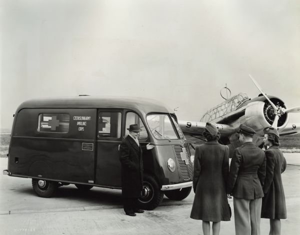 Men and women standing with an International Metro used by Czechoslovakian Relief. An airplane is in the background. All four people are wearing coats and hats. The women's hats include a TWA logo on a ribbon. Original caption reads: "Joseph Simaner of Czechoslovak Relief shows new ambulance unit for service in Britain to TWA Hostesses Eula Walker and Ruth Ellison, and First Lieutenant K.J. Fogle, 108th Observation Squadron." The Metro has a plaque on its side that reads: "Donated by Národní Svaz Ceských Katolíku (National Alliance of Czech Catholics) - America / Federation of American Czechoslovaks of Texas."