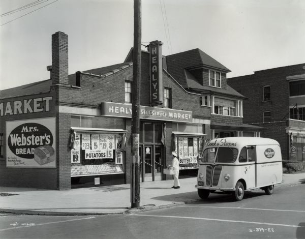 A white International Metro owned by Borck and Stevens bakery is parked in front of the market which is on a street corner. The brick building has two electric signs, and posters with prices filling the windows. A man in a uniform is carrying a large basket of bread up to the door. The brick wall along the sidewalk on the left has a large painted sign for "Mrs. Webster's Bread". Two other buildings are on the right.