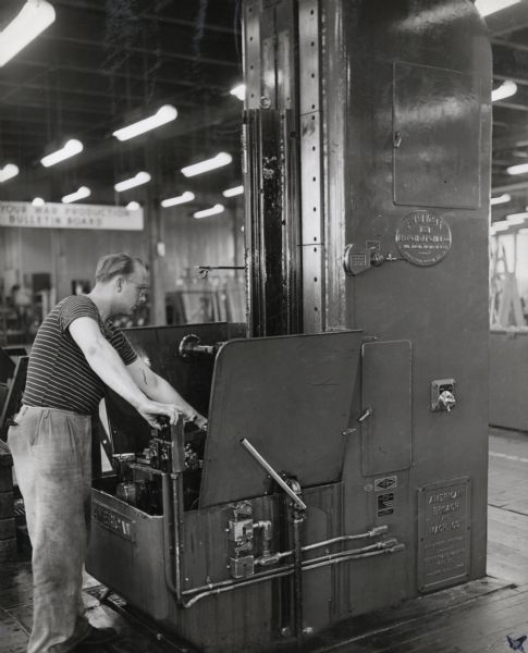 A factory worker operates a vertical broaching machine at International Harvester's 20 (Twenty) millimeter gun plant. Original caption reads: "Vertical broaching is the method followed for making slide grooves and bedding faces on the top surface of the breech block. In this operation an American vertical broaching machine is used. The pieces are fed in one at a time and the ram comes down fast. By this method the pieces hold their size."