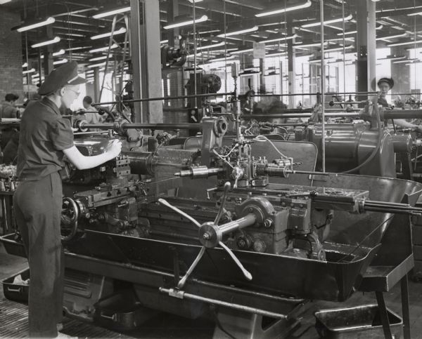 Female factory workers operate lathes at International Harvester's 20 (twenty) millimeter gun plant. Original caption reads: "Women employees have been trained in the Harvester 20 m.m. cannon plant as skillful operators of Jones & Lamson turret lathes. Shown here are six position turrets with a variety of cutting tools. The operations involved on the gun bolt are intricate. Some of the tools are tungsten carbide tip variety. The woman supervisor at the plant is proud of the achievement in training women to operate not only these turret lathes, but broaching, milling and grinding machines as well."