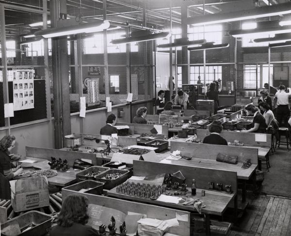 Female factory workers inspect parts at International Harvester's 20 (twenty) millimeter gun plant. Original caption reads: "Many small parts for the gun are made by sub-contractors. These small parts pass through this purchase inspection room on a conveyor and move past the tables of women inspectors. Every such part is inspected by theses Company inspectors and is given a final check by government inspectors."