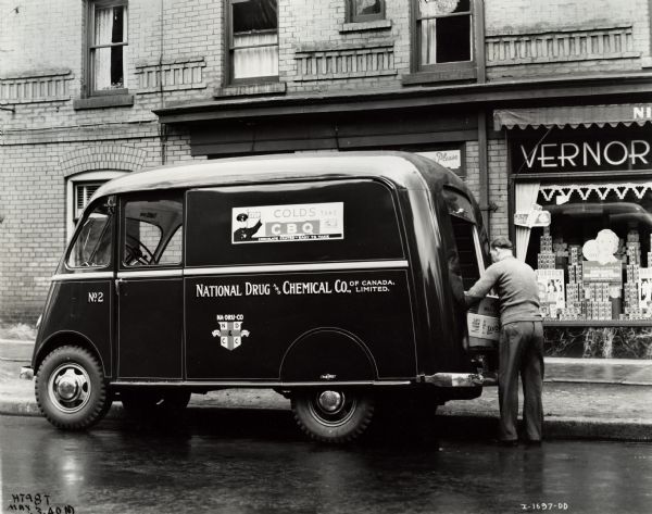 A man standing at the back of an International D-15-M (Metro) truck owned by the National Drug and Chemical Company of Toronto, Canada. The side of the van has a sign for CBQ - a cold remedy. A storefront with products and advertsing are in the background.