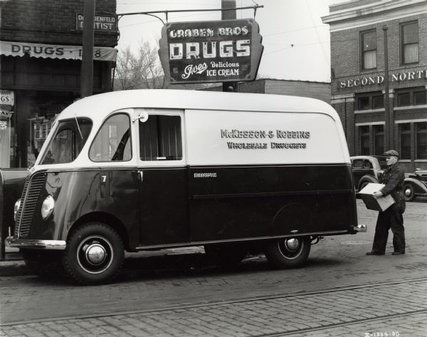 A man lifting a box at the back of an International D-15-M (Metro) truck with 113-inch wheelbase owned by McKesson and Robbins, wholesale druggists. A sign for the Graben Bros. drugstore is above the van.