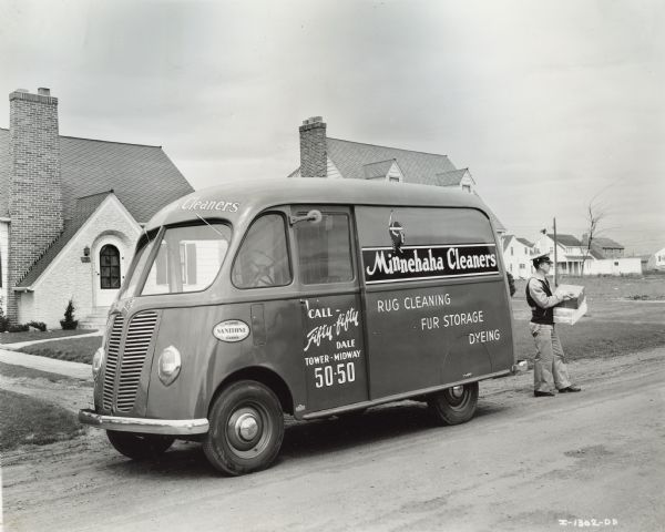 Uniformed man stands with boxes at the back of an International model D-2 truck with 102-inch wheelbase and Metro body. The truck was owned by the Minnehaha Cleaners, and is parked in a residential neighborhood. The logo for the company includes the image of a Native-American woman.