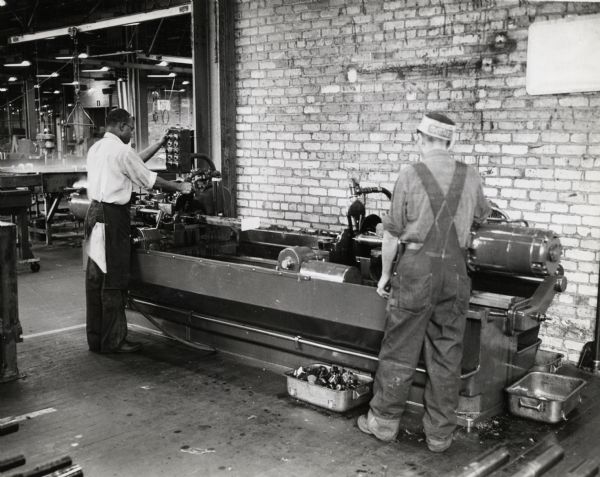 Factory workers at International Harvester's 20 (Twenty) Millimeter Gun Plant operate a factory machine. Caption on photograph reads: "First machining of the bar stock for the barrel, 67.52 inches long when ready for final assembly, is to saw it off at both ends to a length of 70-3/32 inches. The original milling operation for this job has been replaced by sawing with a Sundstrand centering machine and a segmental saw 11 inches in diameter."