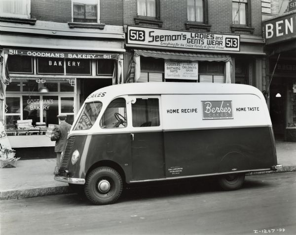 International model D-2-M truck with 115-inch wheelbase and Metro body.  A man in a hat on the left is walking away from the van with some boxes towards Goodman's Bakery. To the right of this storefront is Seeman's Ladies and Gents Wear.